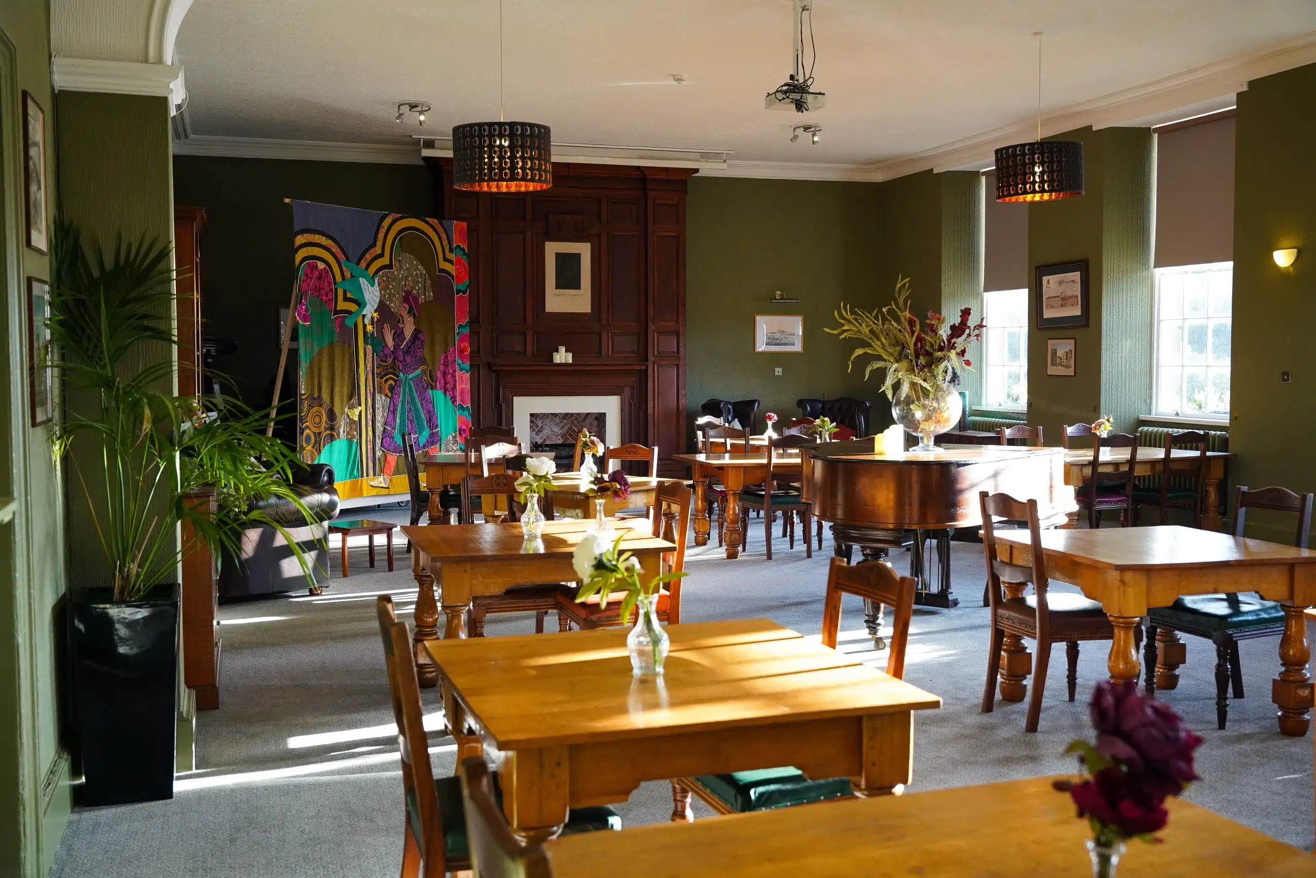 Cosy room with wooden tables, floral vases, a grand piano, and natural light from sash windows