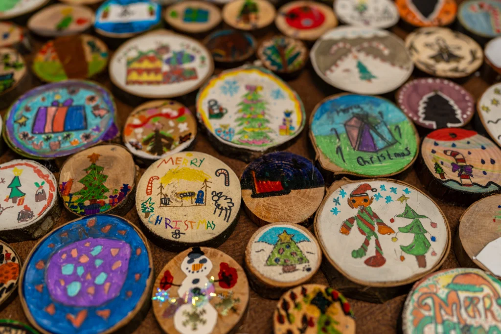 A collection of small, round wooden ornaments decorated with hand-painted Christmas drawings by local schoolchildren, featuring festive designs like snowmen, Christmas trees, and "Merry Christmas" lettering.